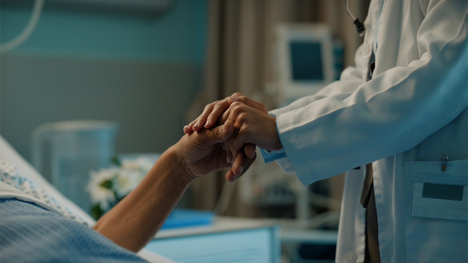 Clinician holding the hand of a patient lying in a hospital bed. No faces are seen.